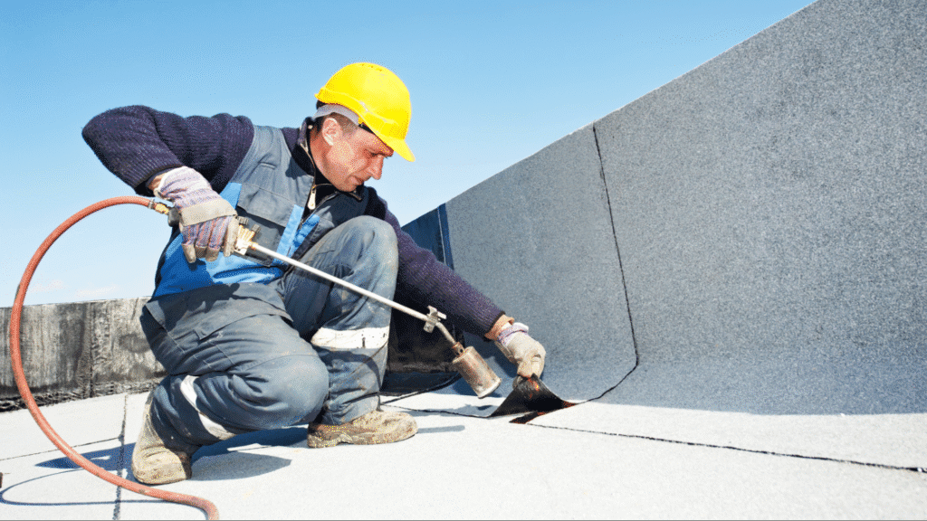 A commercial roofing contractor uses a heat-welding torch to fuse a grey TPO membrane seam on a flat roof.